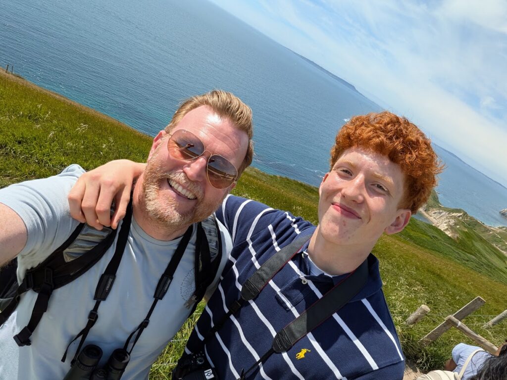 Two people stand at a coastal cliff location looking into the camera with view of the sea behind them